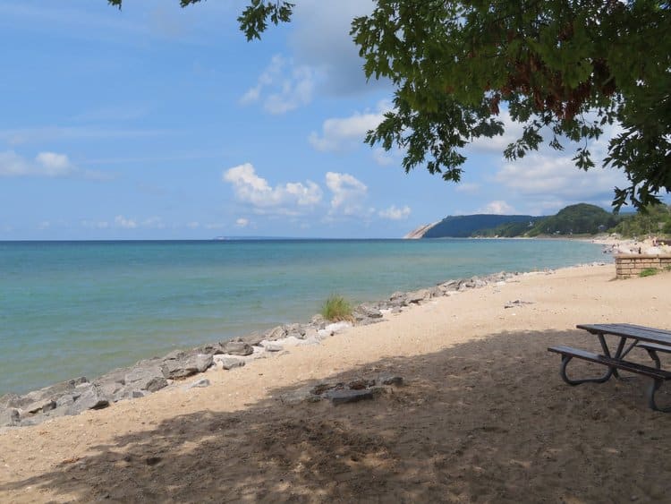 a sandy beach with a picnic table on it lines the shores of Lake Michigan at Sleeping Bear Dunes National Lakeshore.  Using a beach wheelchair to go to the beach is one of the things to do in Sleeping Bear Dunes.