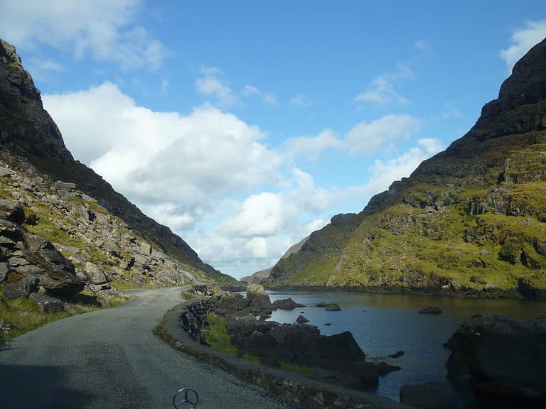 A narrow road winds between two mountains.  A stream is to the right of the road in Ireland.