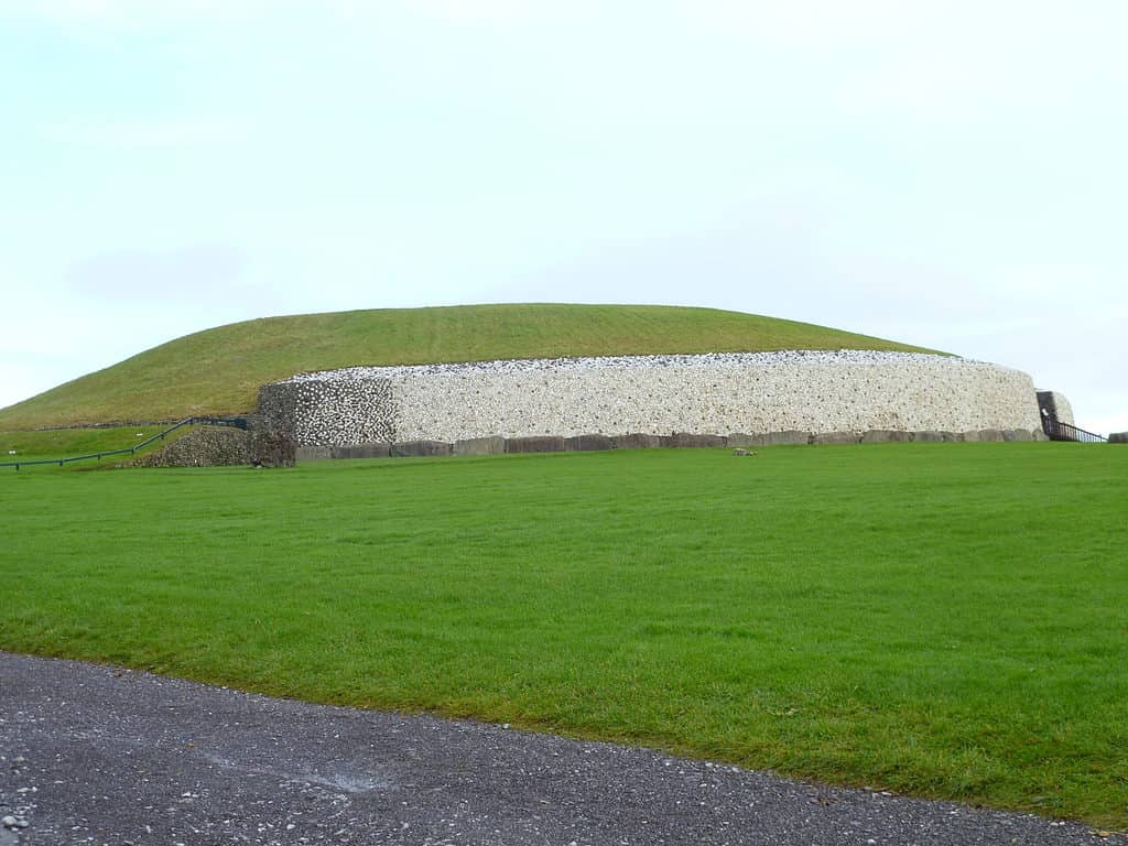 A green mound in a field of green grass has stone sides that make up its circumference.  Newgrange is one of the cultural sites in Ireland is a must visit according to my Ireland travel tips