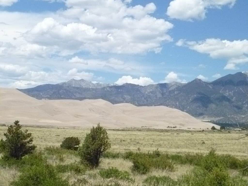 A large sand dune collection in a grassy field at the base of a mountain range (located on the right)