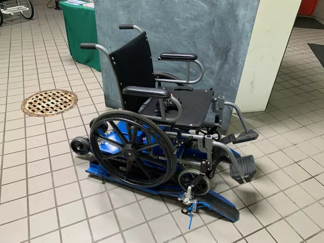 a black manual wheelchair sitting on a set of tank tracks that attaches to the wheelchair to make it adapted for hiking