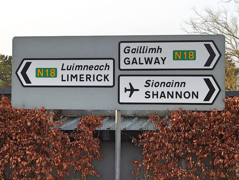 A large gray road sign has three smaller white directional signs on it.  The white directional signs have Gaelic and English wording.  Ireland travel tips: road signs are in both Gaelic and English