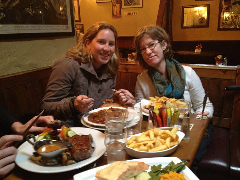 Two blonde-haired white women are sitting in a booth in a restaurant with plates of food in front of them.  