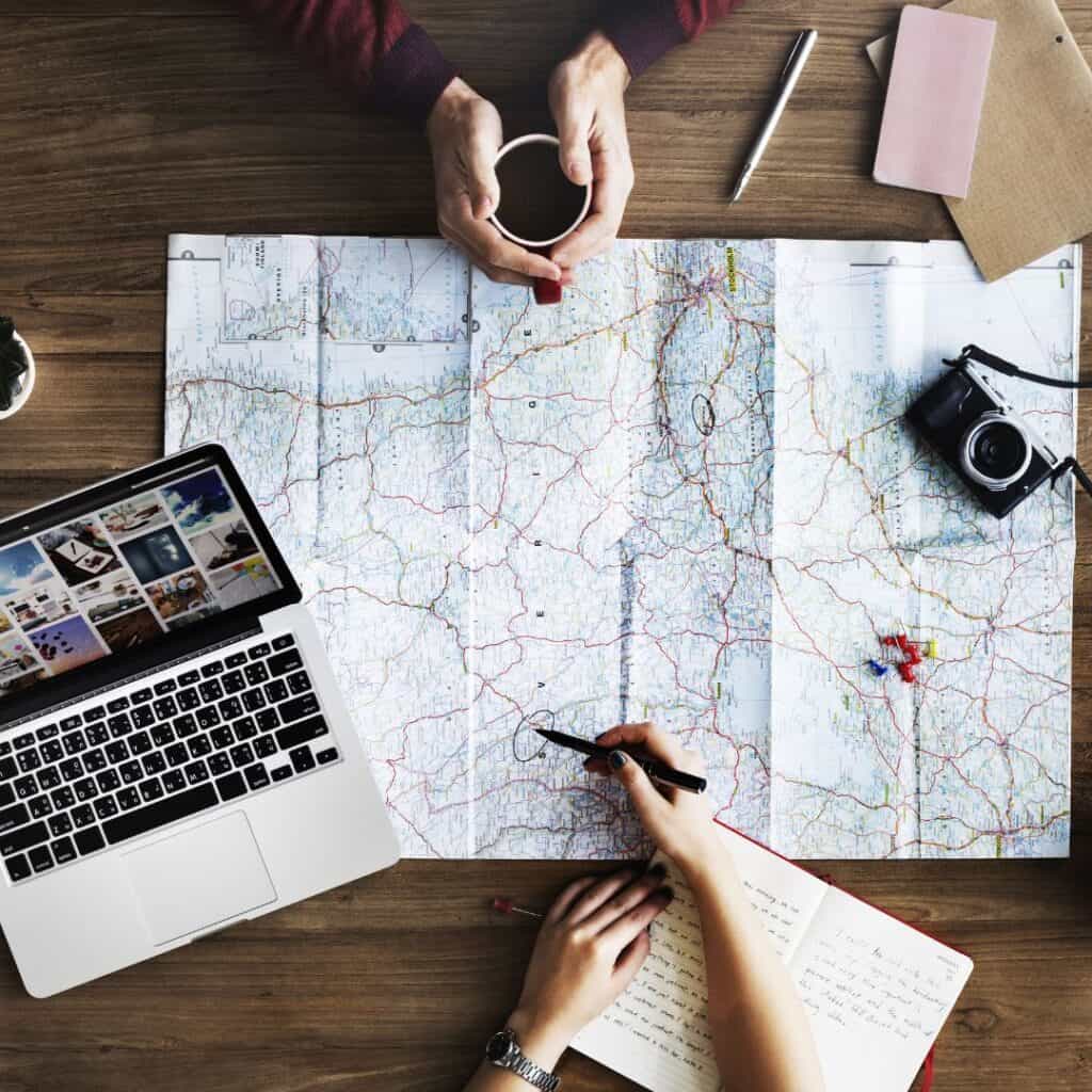 a white map lays on a wooden surface and is surrounded by a black camera on the right, an open laptop on the left and an open journal with a white arm holding a pen over it on the bottom