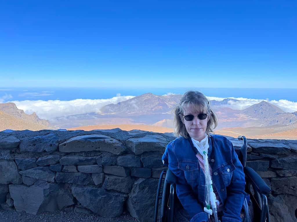 I'm sitting in a wheelchair with a jean jacket and am in front of the crater at Haleakala National Park.  The crater is brown with reds and oranges in the soil.  There is a clear blue sky overhead.