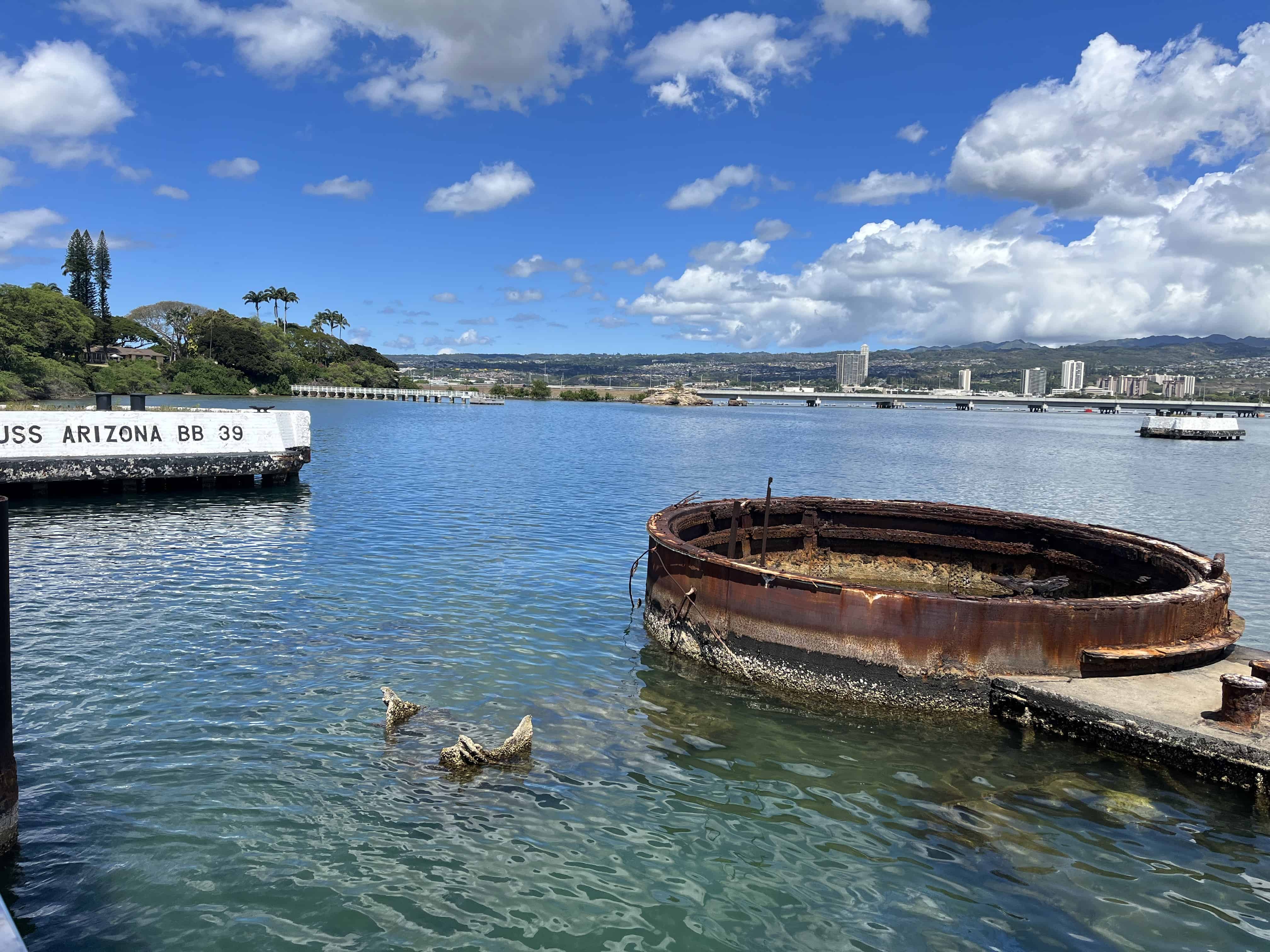 On the bottom right, a rusted cylinder emerges from the water. On the lefthand side of the picture a white rectangular buoy has black letters on it saying USS Arizona