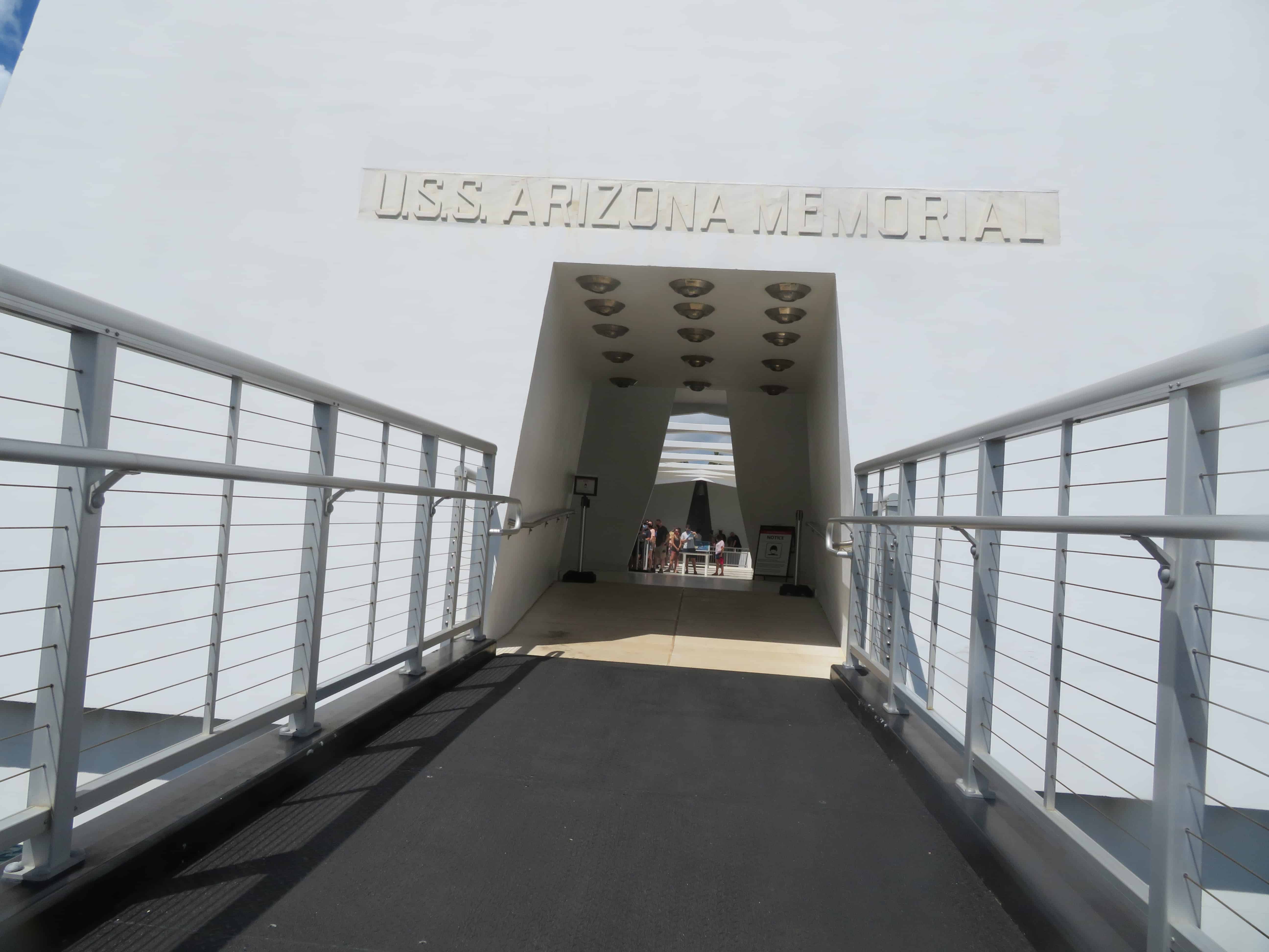 a black ramp leads to a white building with an open doorway.  Over the door is an off-white set of words that say USS Arizona Memorial