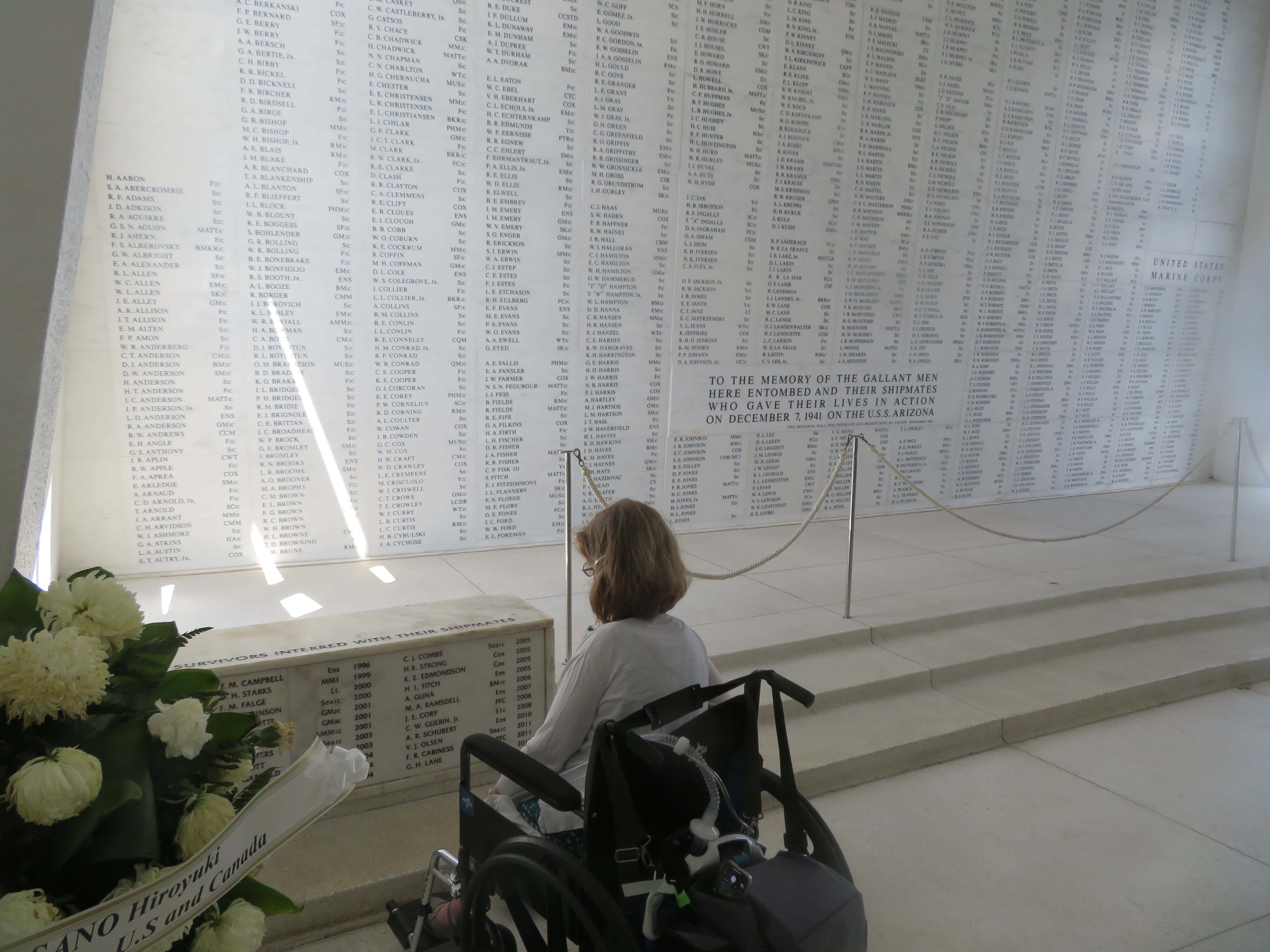 A white wall with black names in columns covering the whole wall are in the background. In the foreground, on the left are white flowers. In the center I'm sitting in a manual wheelchair and looking at the wall.