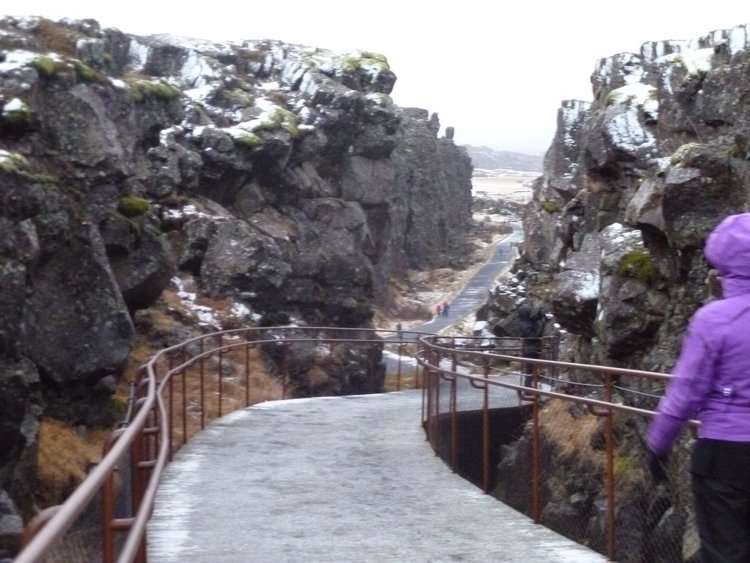 A paved pathway with rails on either side passes between two black rocky outcroppings in Pingvellir National Park in Iceland