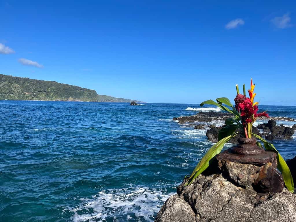 A large rock is on the right overlooking the ocean on Maui.  The rock has a bouquet of red and orange flowers on it.