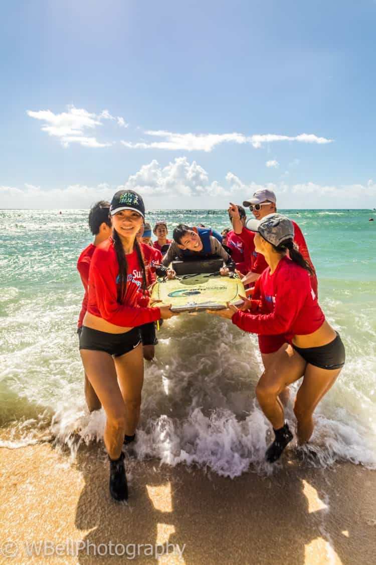 6 people in red jackets carry a yellow surfboard out of the ocean with a male laying on the surfboard.