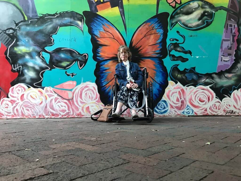 along a grey sidewalk is a colorful mural on the side of a building featuring a blue and orange butterfly.  I (a white woman in a blue skirt and a blue jean jacket) am sitting in a manual wheelchair in front of the butterfly