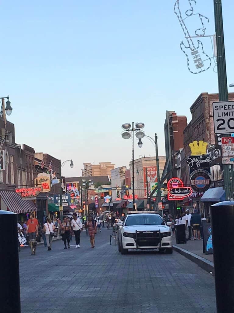 Beale Street in Memphis Tennesse has a white police car on the right, and people walking in the street which is lined with buildings with neon signs