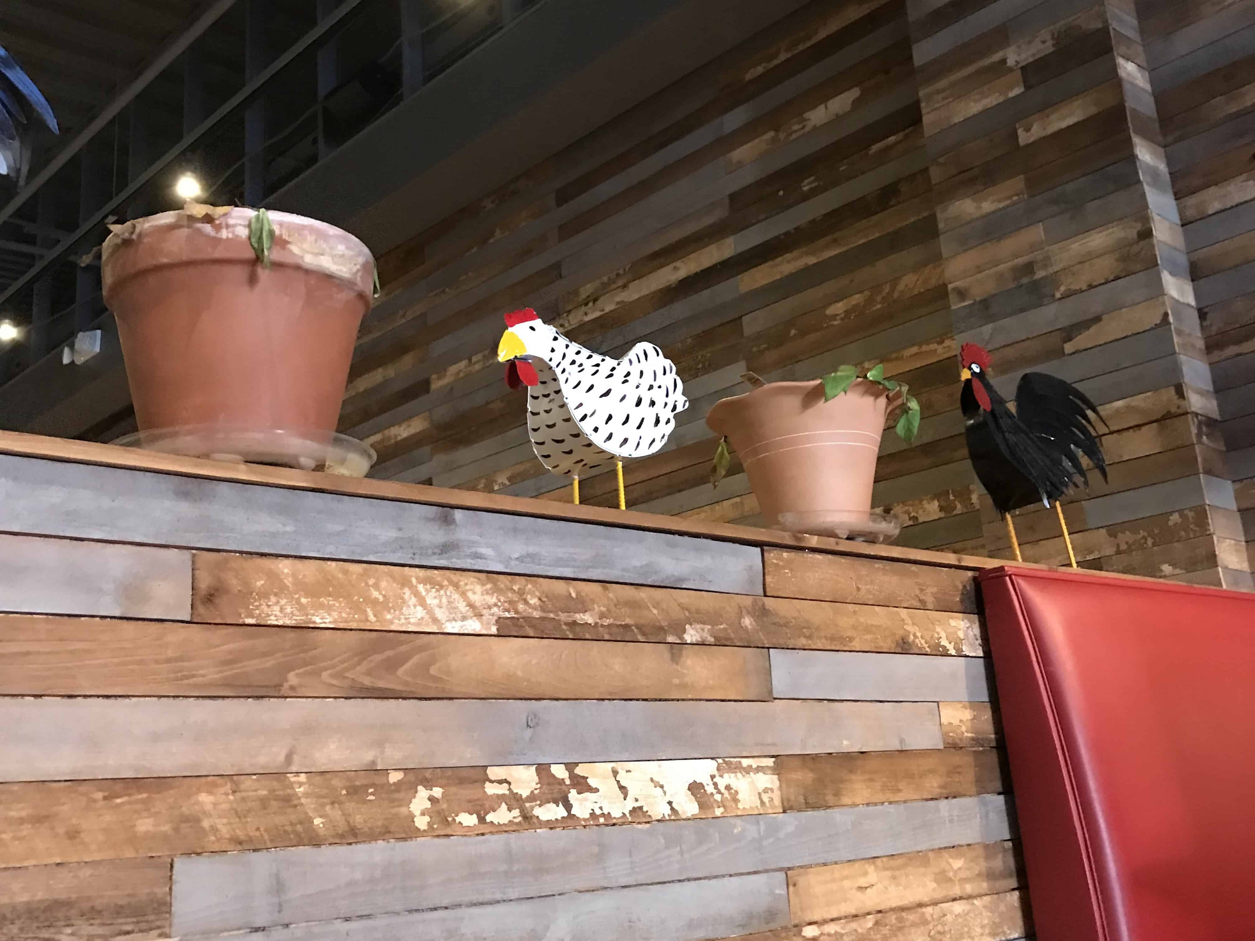 The inside of the restaurant Sugar Grits has a multi colored grey and brown wall with two brown flower pots sitting on top.  In the middle of the pots is a white chicken