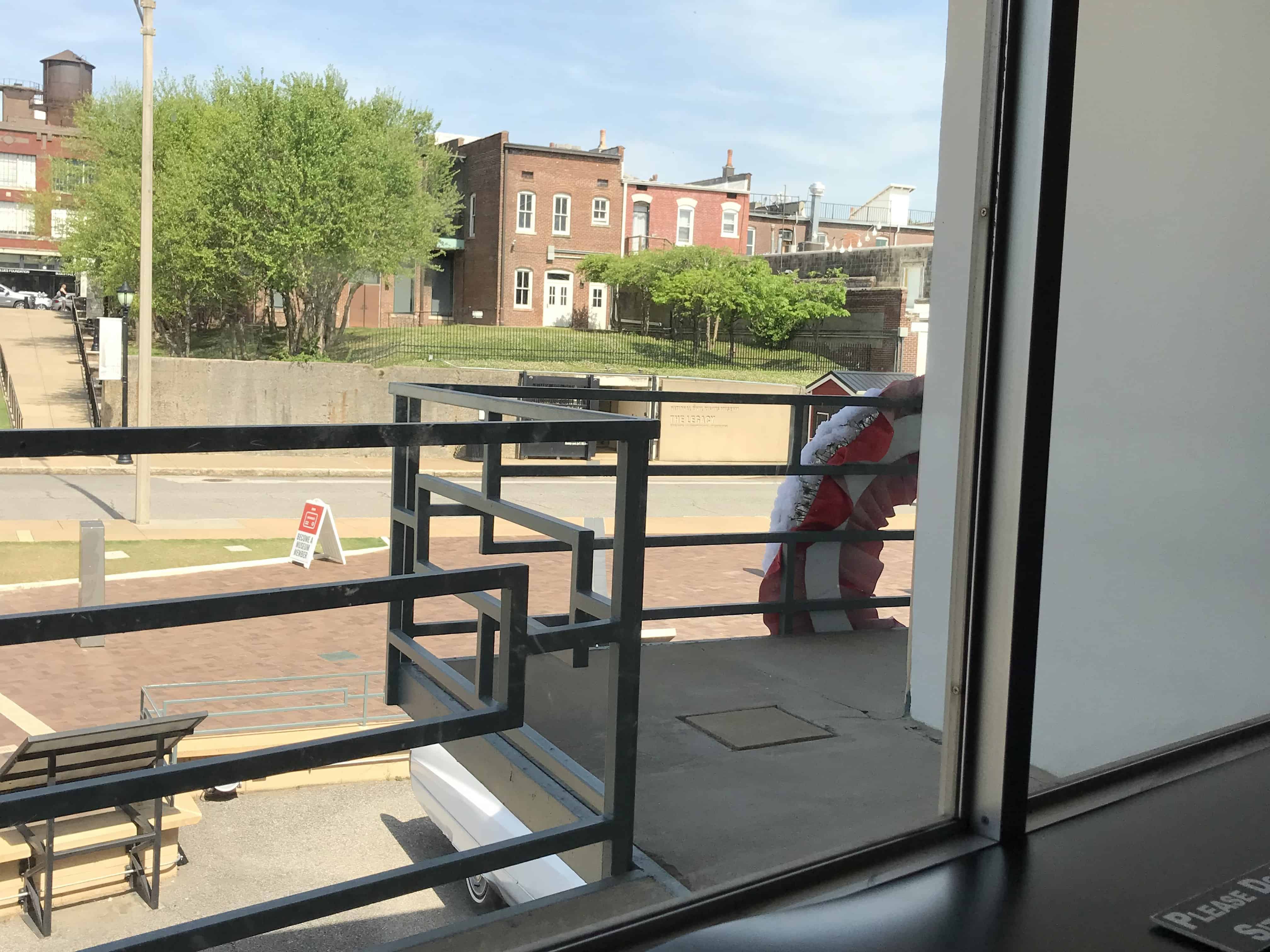 Looking out at the balcony where Martin Luther King Jr was assassinated as seen from inside the Civil Rights Museum. A white wall is on the right with a dark floored balcony on the bottom. An iron rail with a flowered wreath on it runs across the middle of the picture