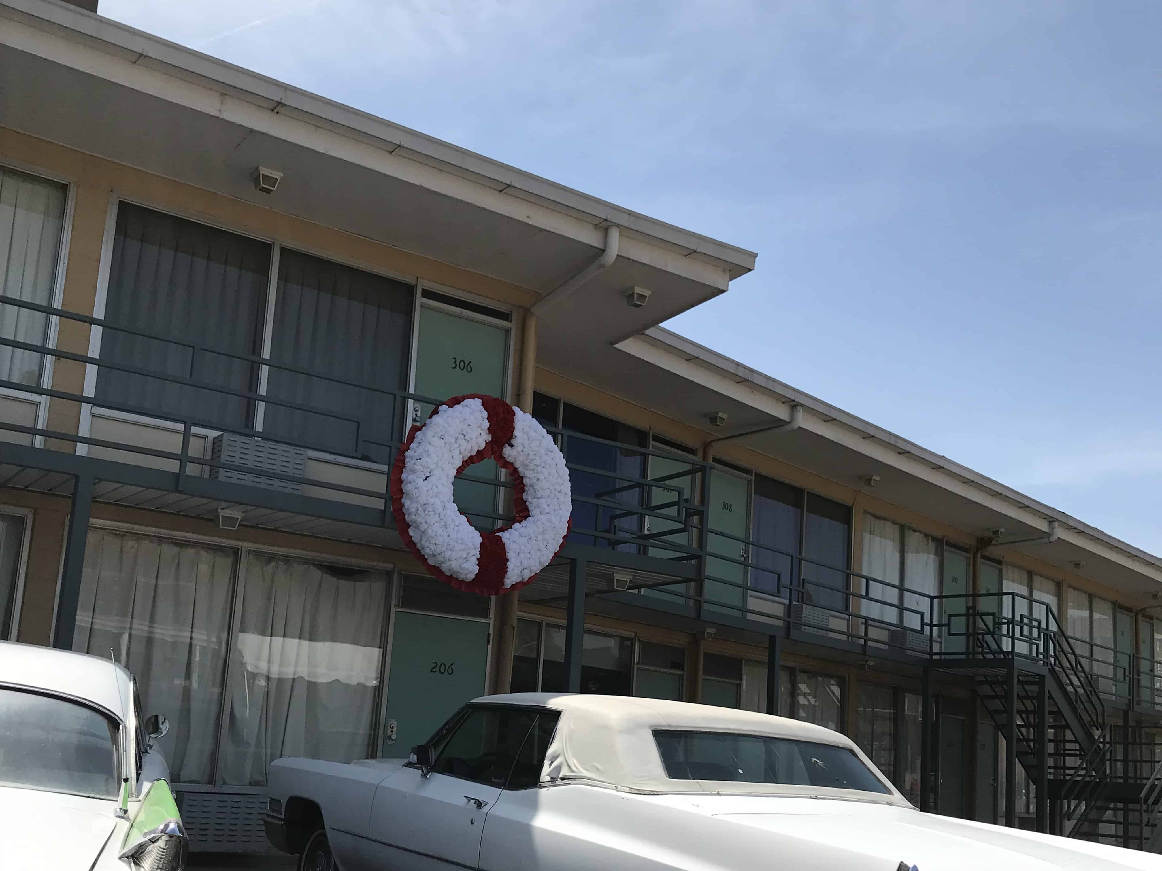 a white car from the 1960's sits in front of the Lorraine Motel. The second floor of the hotel has a balcony with a flowered wreath hanging on the iron railing