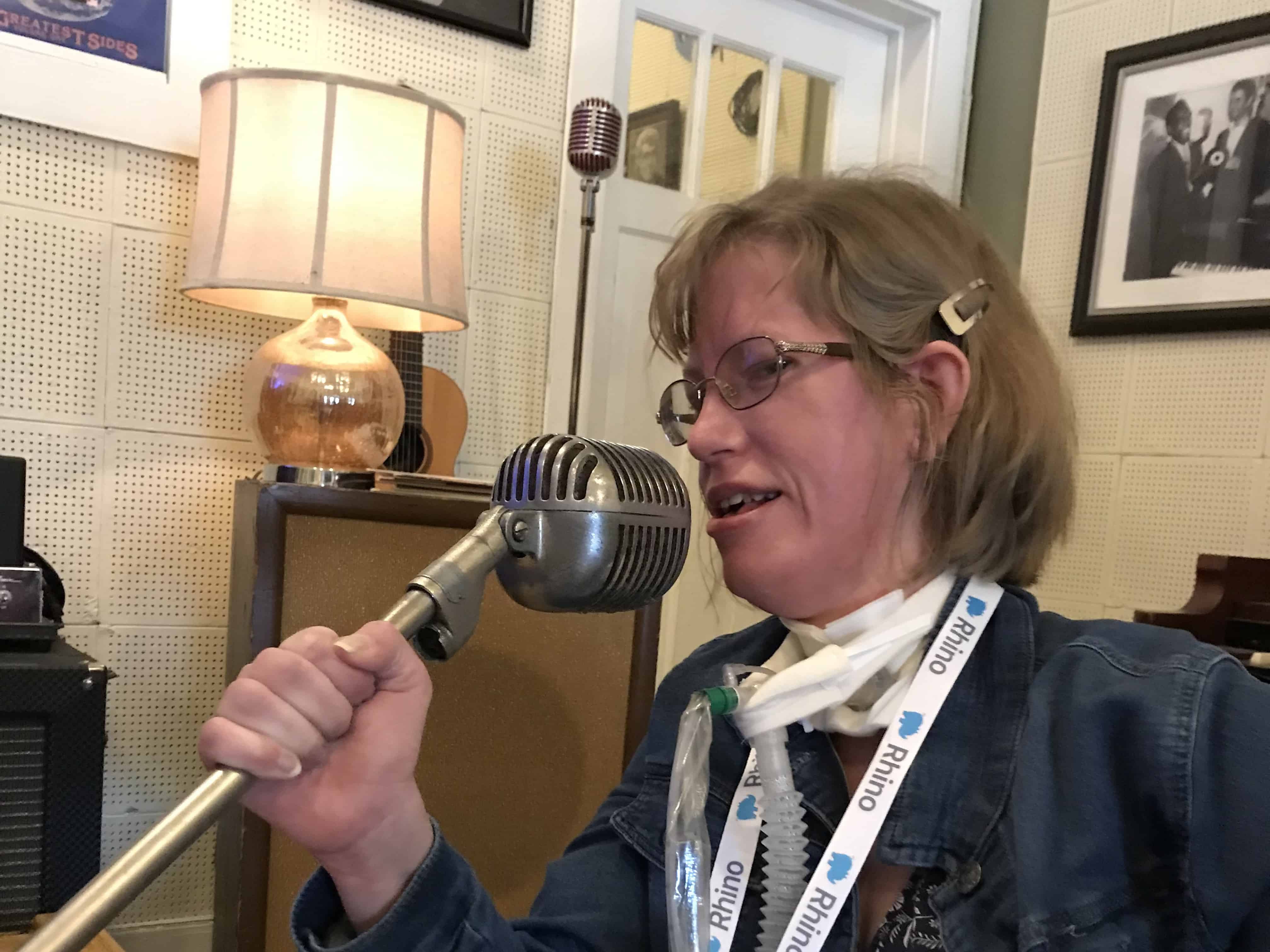 a white woman with blonde hair (me) is wearing a blue jean jacket and holding a microphone on a stand at an angle and singing into it.