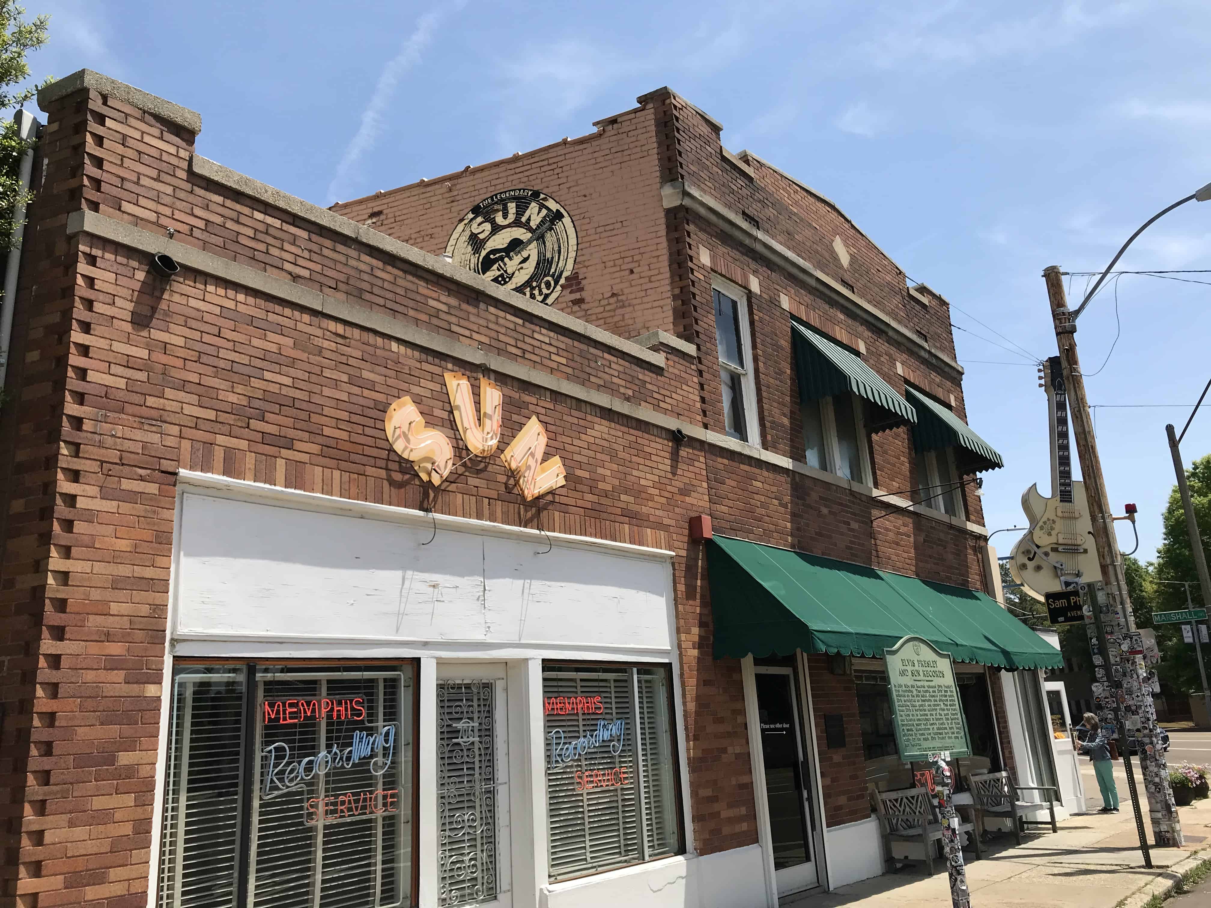 a brick two story building with a green awning. To the left of the building is a one story structure attached to the building. The structure has white trim and a red sign above the door that says "sun"