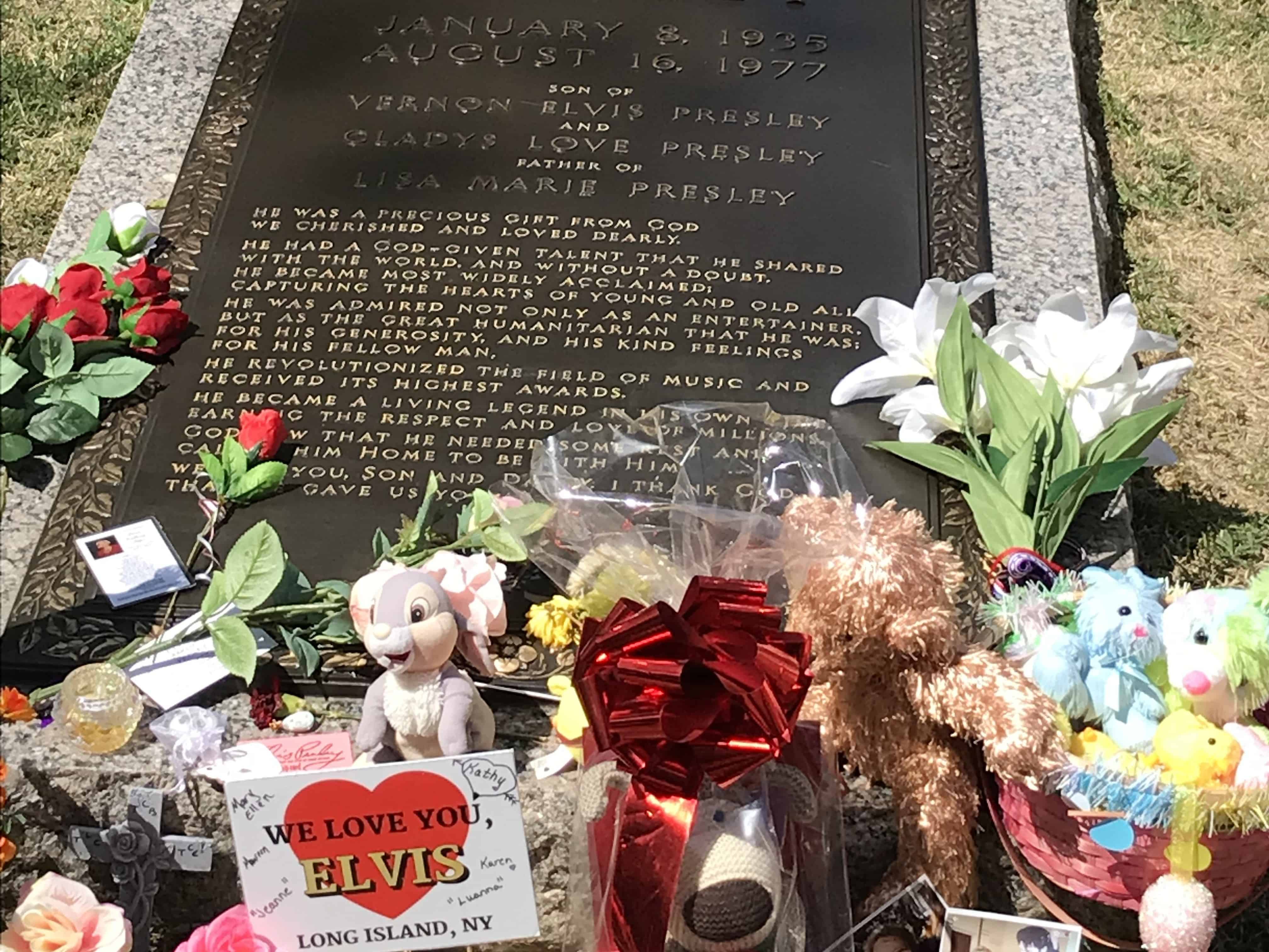 A grey stone with a dark brown plaque makes up the grave of Elvis Presley. Flowers and gifts are laid at the base of the grave that lies flat on the ground