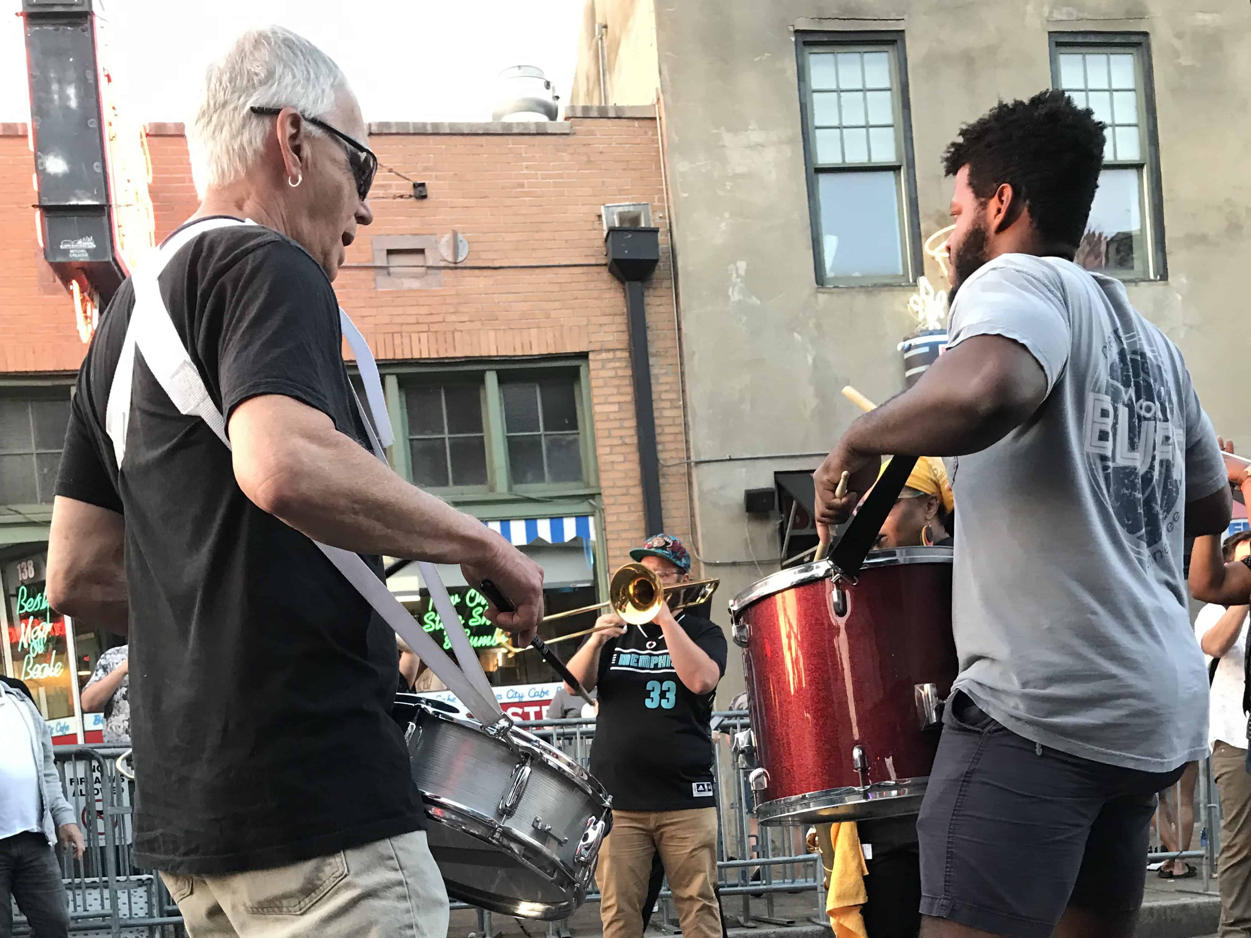 A circle of male musicians stand on Beale Street in Memphis playing drums, saxophone, and trumpet