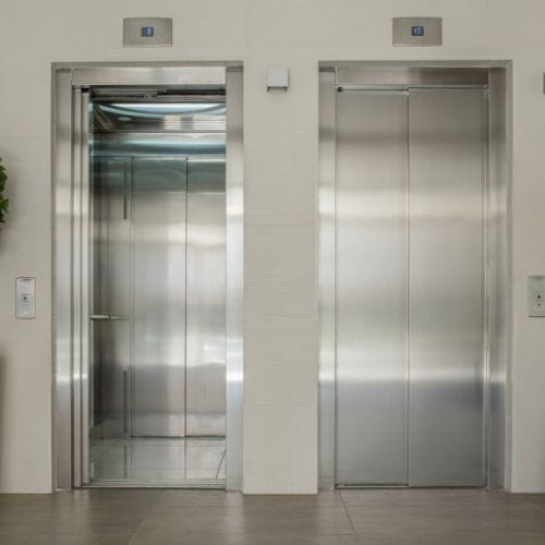 two silver elevators are side by side on a beige wall.  The elevator on the left has it's doors open.