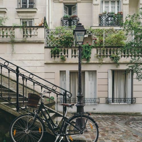 a cobblestoned road leads to a staircase with a bike in front of it.  In the background is a white building with wrought iron windows and green plants hanging from the windowsills.  Cobblestones can be one of the biggest challenges during a wheelchair accessible cruise to Europe 