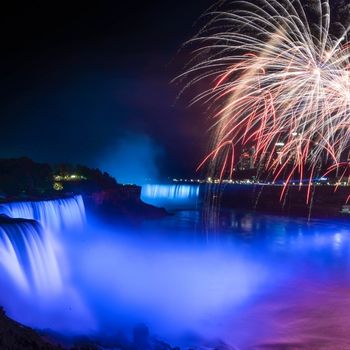 Niagara Falls is lit up at night with blue lights and red fireworks go off in the sky in the upper right corner