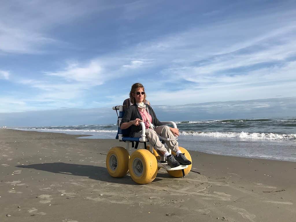 a white woman with blonde hair is sitting in a blue beach wheelchair with large yellow wheels