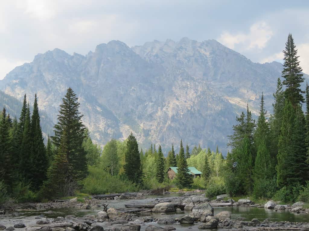 a rocky stream leads to tall pine trees at the base of the Teton Mountains