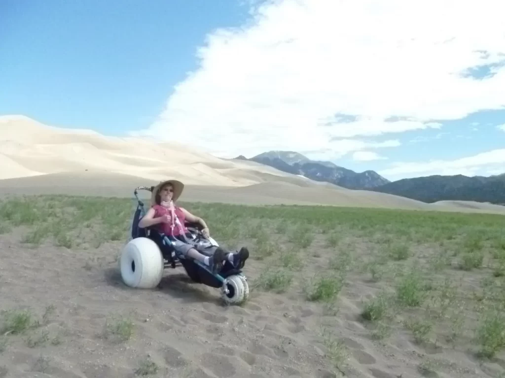 a white woman in a large sun hat is sitting in a beach wheelchair in front of a large sand dune with mountains on the right in Great Sand Dunes National Park