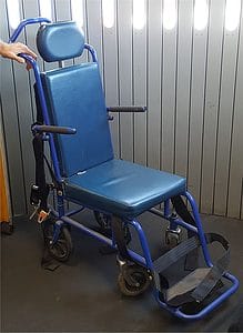 A blue, high backed aisle wheelchair with a head rest sitting in front of a wall at an airport