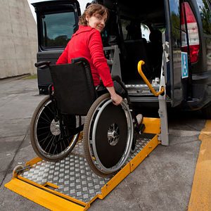 a white woman with a red shirt is in a manual wheelchair and using a lift at the back of a van to enter it.