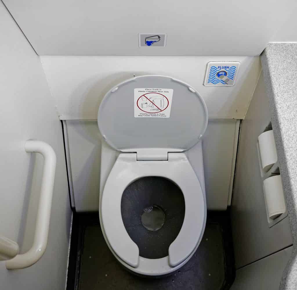 a narrow restroom on an airplane has an open toilet in the middle, a white grab bar on the left side of the photo and a counter with a sink on the right side of the photo