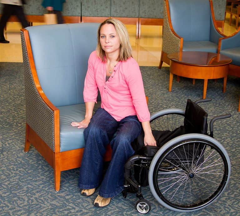 a white, blonde-haired woman in a pink shirt and jeans is self transferring from her manual wheelchair to a blue chair