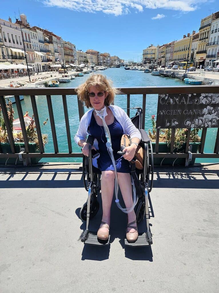 A blonde-haired white woman in a blue dress sits in a manual wheelchair in front of a canal in Sete France