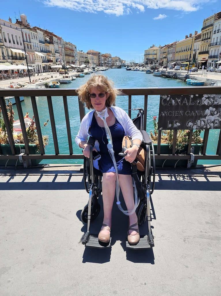 A blonde-haired white woman in a blue dress sits in a manual wheelchair in front of a canal in Sete France