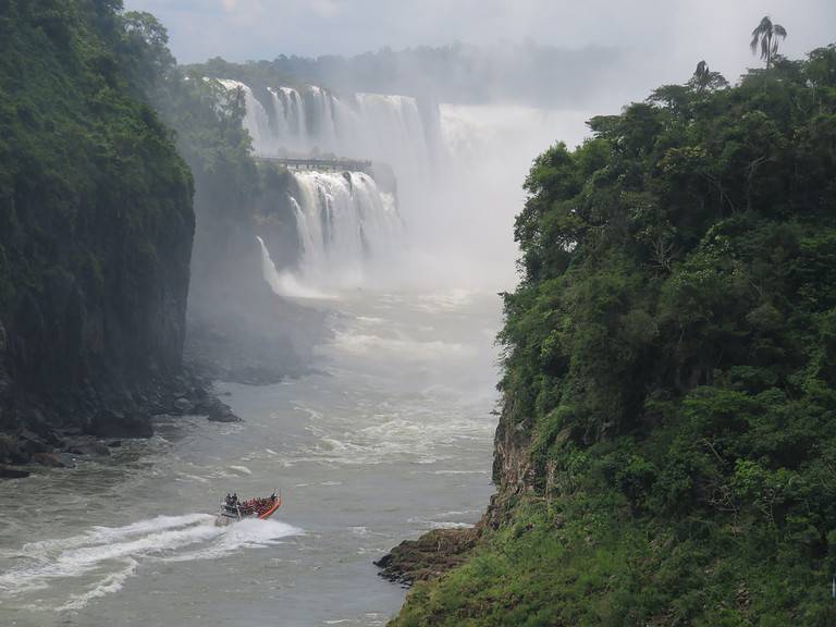 An orange boat is going up river towards a large U shaped waterfall that makes up Devil's Throat at Iguazu Falls