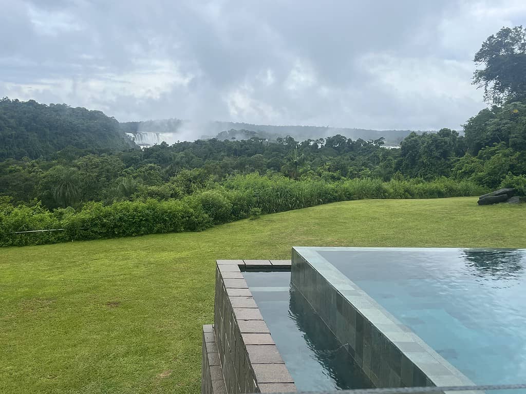 A blue and gray infinity pool sits on a lush green lawn in front of green trees and Iguazu Falls at the Gran Melia Iguazu hotel