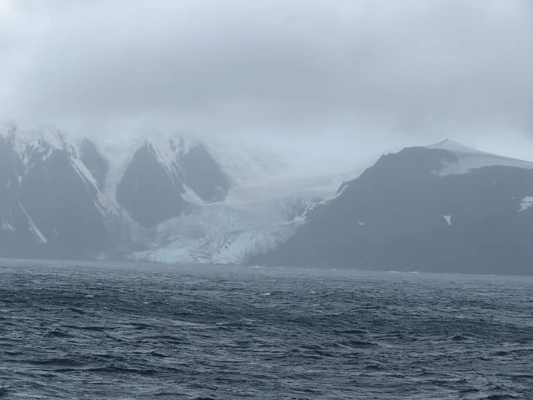 A blue glacier slides down to the water between two snow-covered mountains in Antarctica