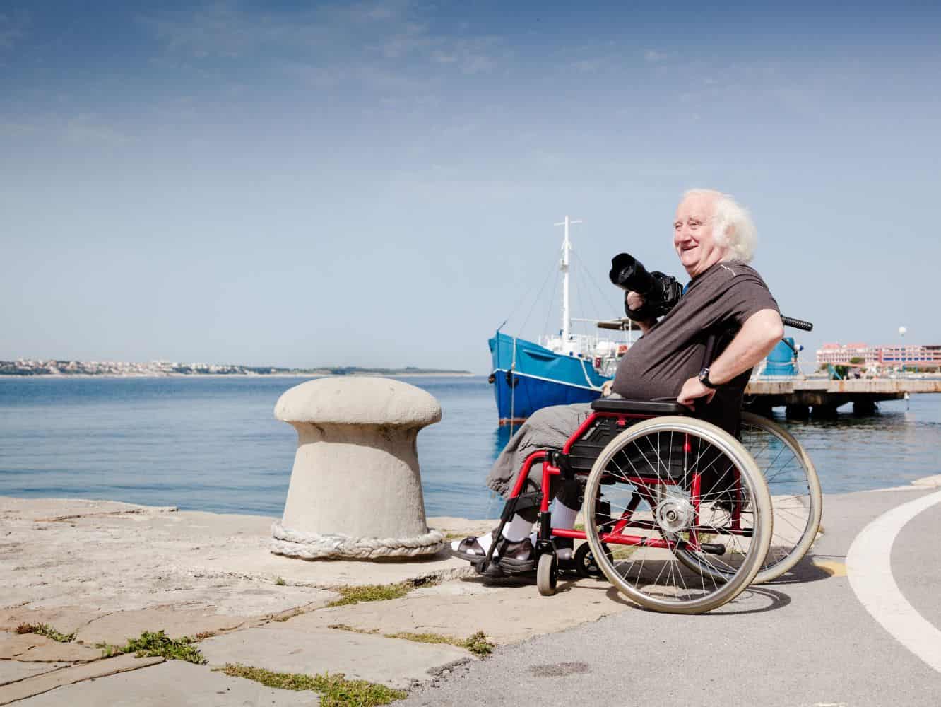 An older man with white hair sits in a wheelchair holding a large black camera near the waterfront.