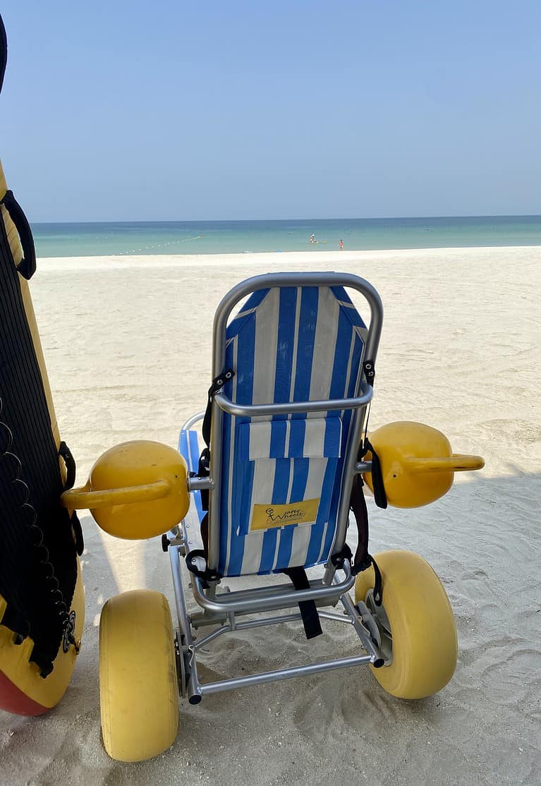 a beach wheelchair with big yellow tires and a blue and white seat sits on a white sand beach in Sharjah UAE