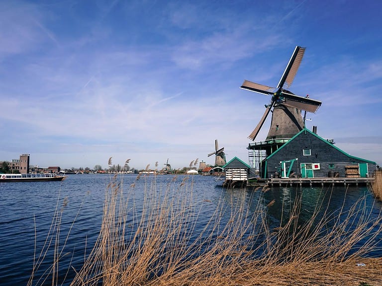 a series of windmills with dark buildings and green trim sit alongside a river just outside of Amsterdam and make of Zaanse Schans.