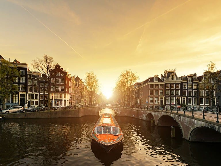 The sun sets over a canal in Amsterdam which has an orange canal boat cruising down it.