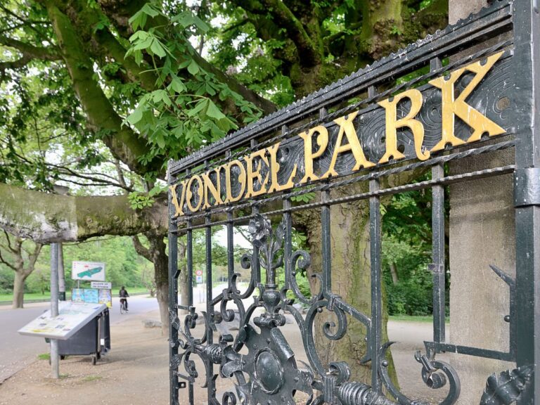 A dark iron gate with gold lettering that says "VondelPark" leads to paved pathways lined with trees.