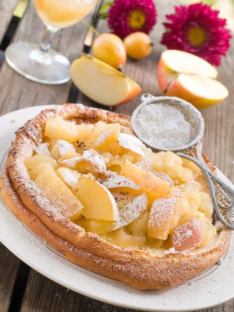 A dutch pancake topped with apples sits on a white plate and is dusted with powdered sugar.