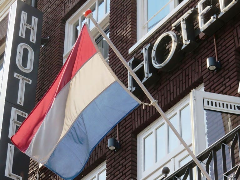 A brown brick building with while windows has the word "hotel" on it with the flag of the Netherlands hanging from an iron balcony.