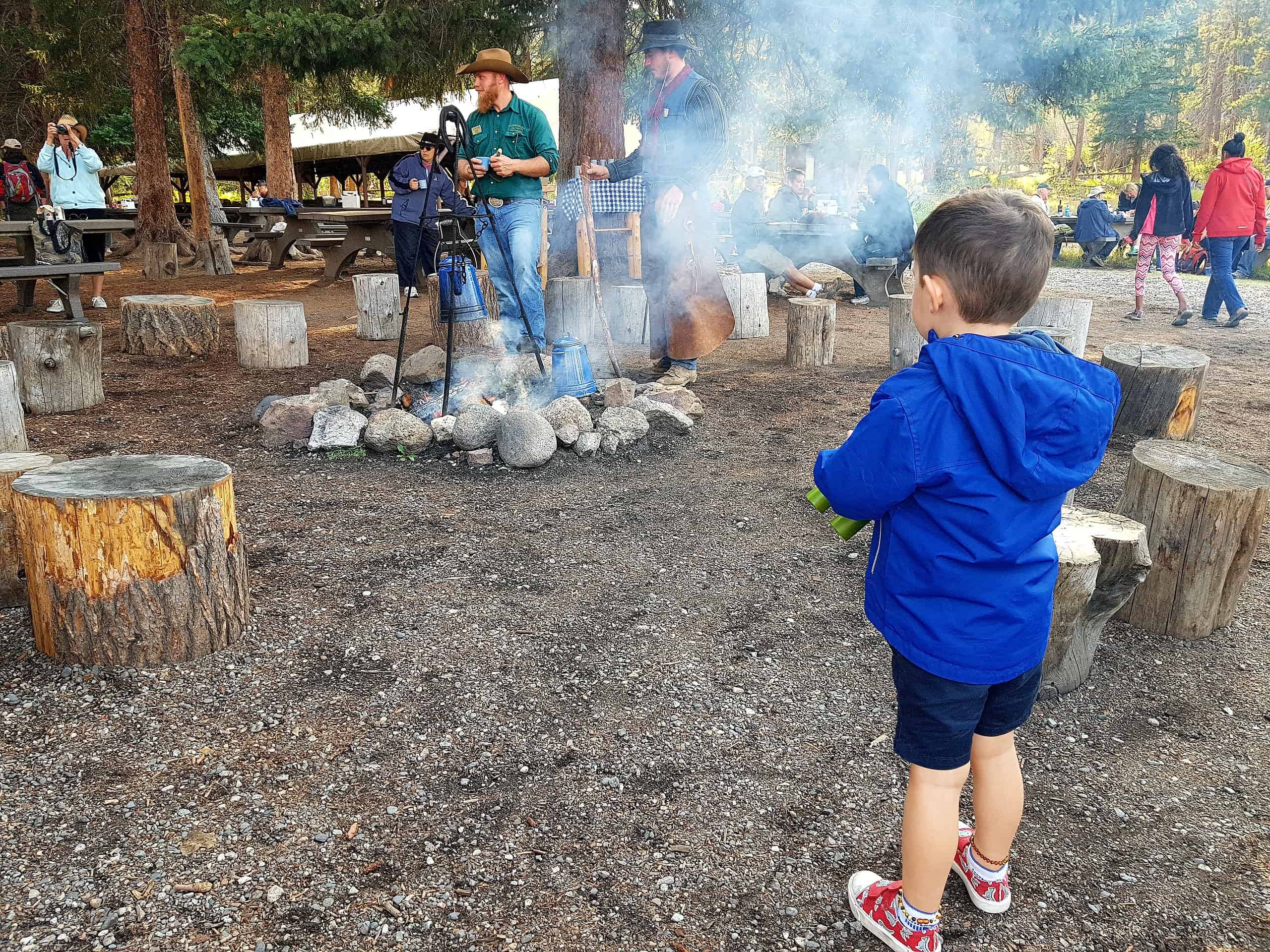 A young white boy with dark hair is wearing a blue jacket and dark shorts and watching adults cook over a campfire.