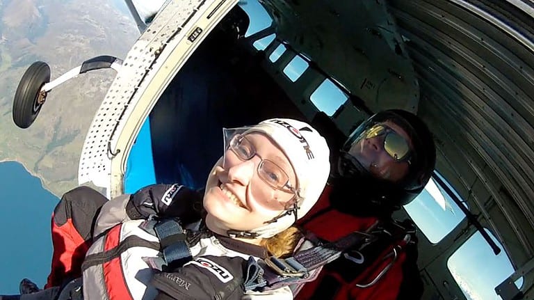 A white woman is sitting in an open door of an airplane high above the earth as she prepares to skydive.  Traveling the world changed her personality by allowing her to become more confident.