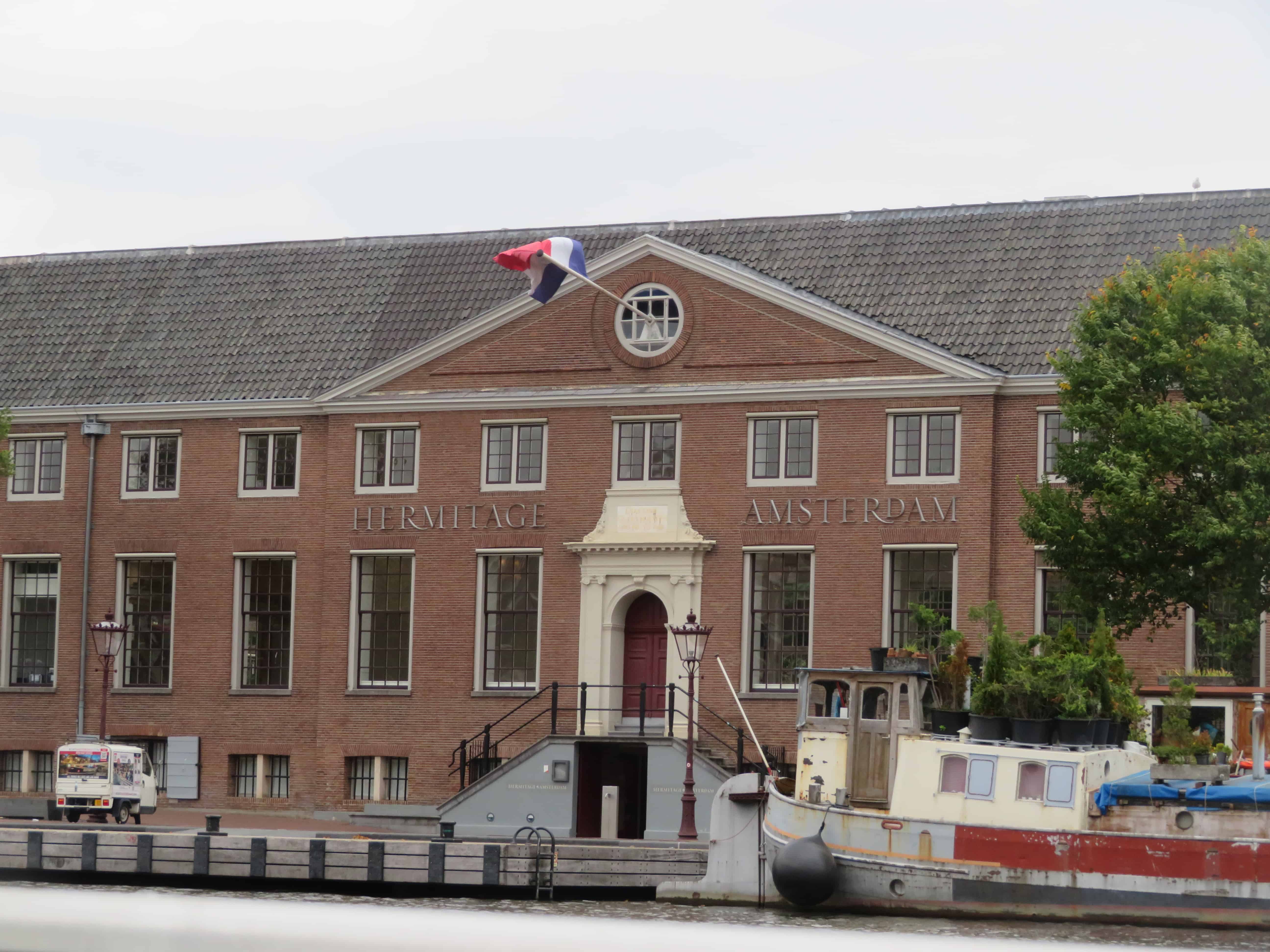 A large brick building with grey roof and cream colored ornate doorway is the Hermitage Museum in Amsterdam.