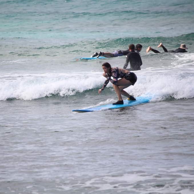 A woman surfing in shallow water on a blue surfboard.  Trying new things when you travel, such as surfing, can change your personality and make you braver.